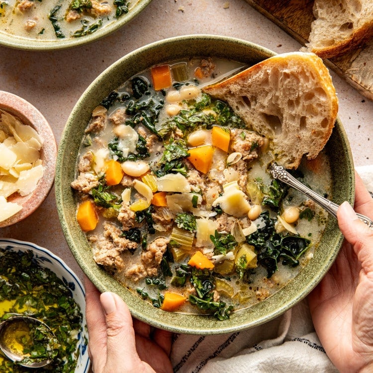 Sausage, kale and white bean soup in bowl with a side of crusty bread and spoon in bowl.