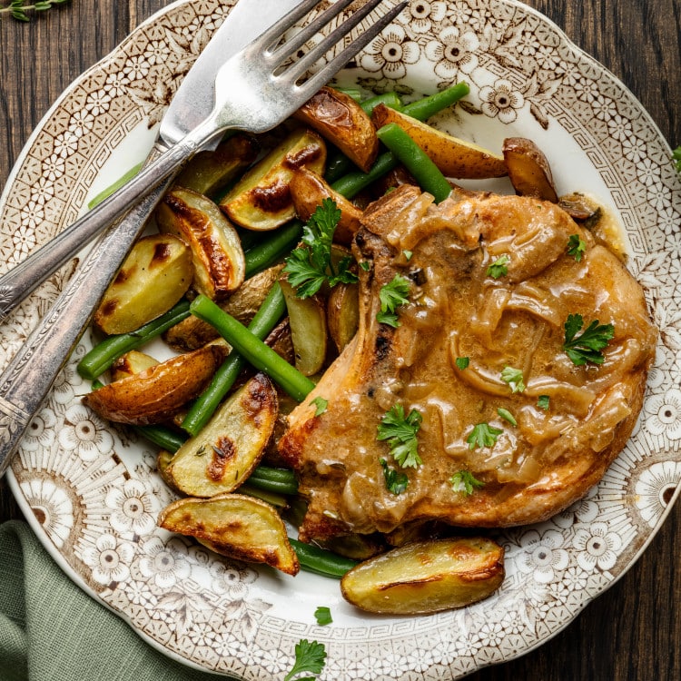 Slow cooker pork chop on plate with potatoes and green beans with knife and fork.