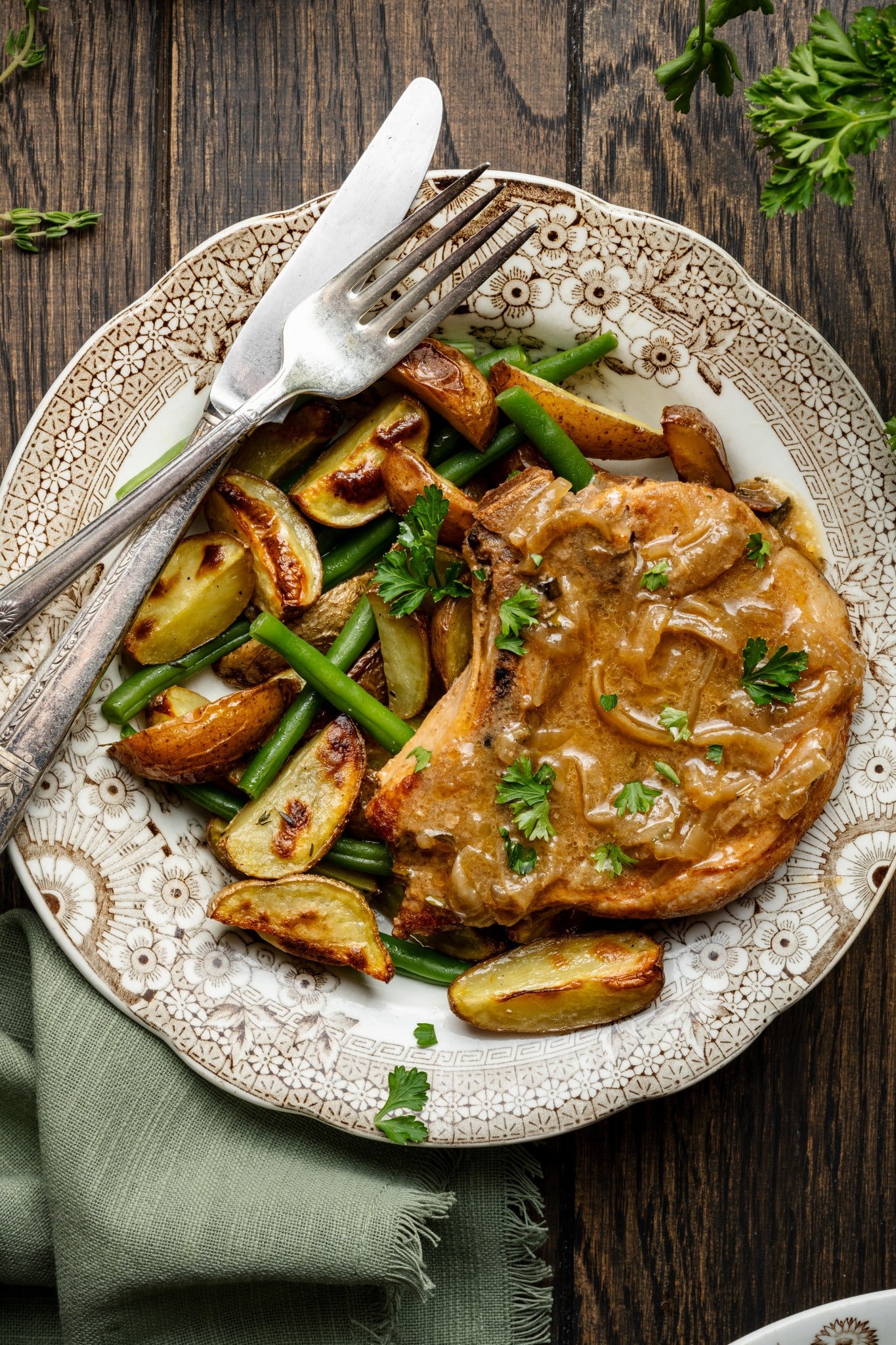 Slow cooker pork chop on plate with potatoes and green beans with knife and fork.