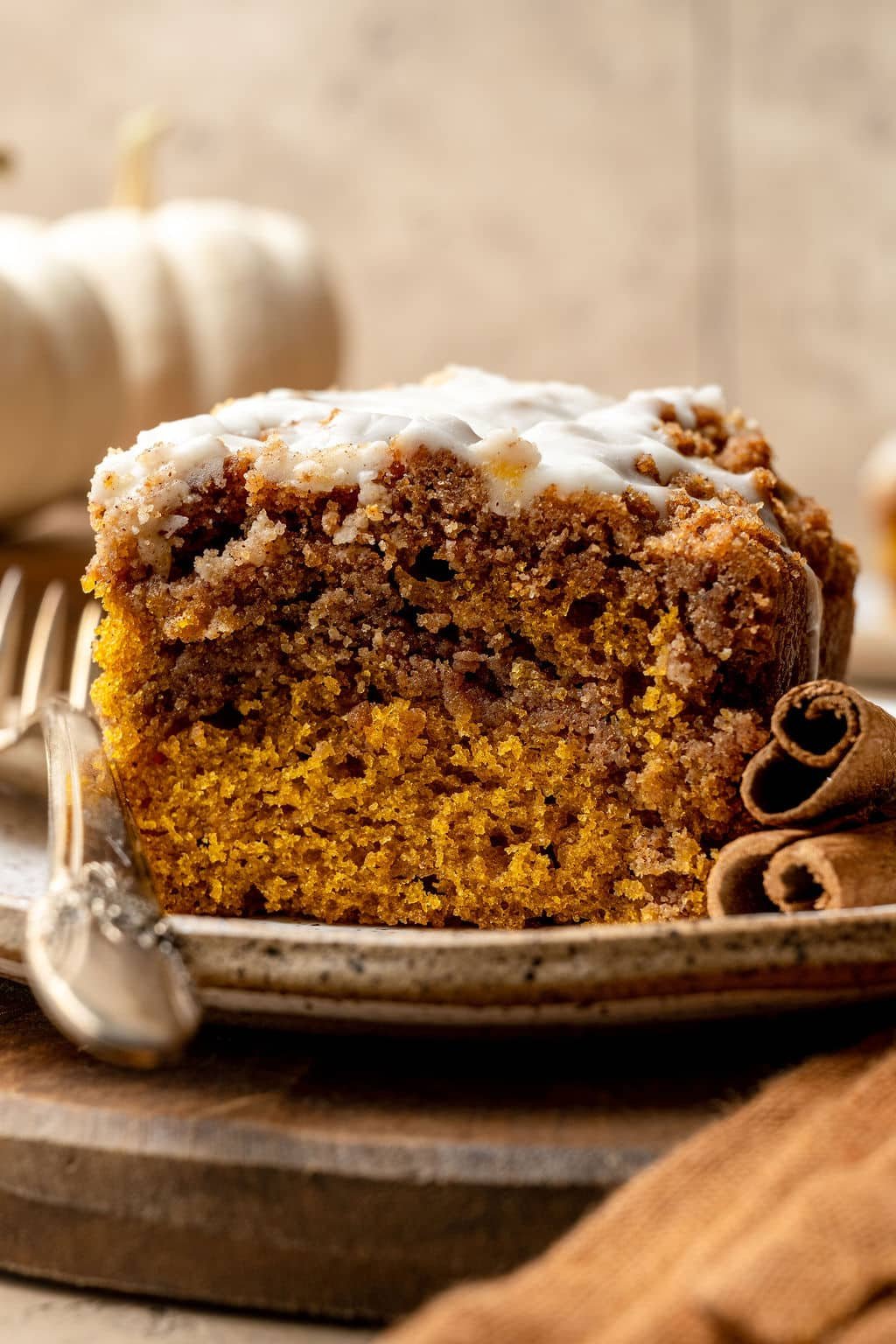 Close-up of a slice of pumpkin coffee cake with a cinnamon swirl and white glaze, served on a speckled plate with a silver spoon. Cinnamon sticks rest beside the plate and small white pumpkins are in the background.