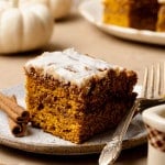 A slice of pumpkin coffee cake with cinnamon swirl and glaze on a plate with a fork, styled with cinnamon sticks and white pumpkins in the background.