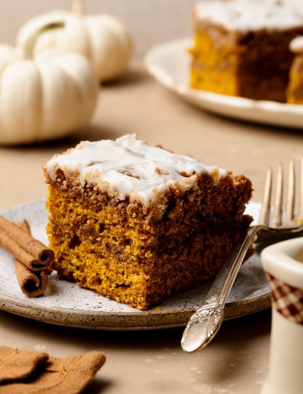 A slice of pumpkin coffee cake with cinnamon swirl and glaze on a plate with a fork, styled with cinnamon sticks and white pumpkins in the background.