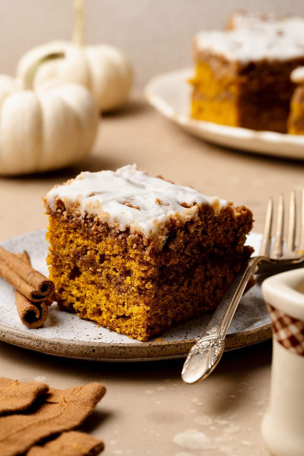 A slice of pumpkin coffee cake with cinnamon swirl and glaze on a plate with a fork, styled with cinnamon sticks and white pumpkins in the background.