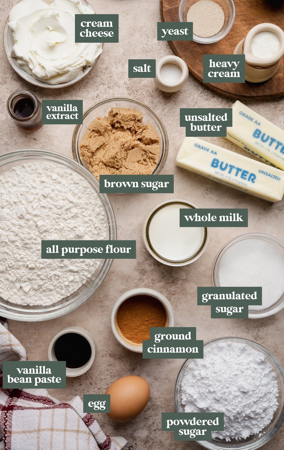 Overhead image showing labeled ingredients for small batch cinnamon rolls arranged on a light beige surface. Ingredients include bowls of all-purpose flour, brown sugar, powdered sugar, granulated sugar, ground cinnamon, cream cheese, vanilla extract, vanilla bean paste, an egg, yeast, salt, whole milk, heavy cream, and sticks of unsalted butter. Each ingredient is neatly labeled with green text boxes, creating a clear and organized ingredient layout.