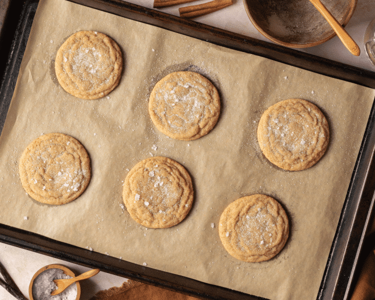 Six brown sugar cookies baked and sprinkled with flaky sea salt on parchment-lined baking tray.