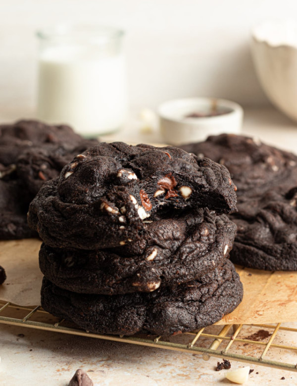 Three black and white chocolate chip cookies stacked on top of each other on wire rack with bite taken out of top cookie.