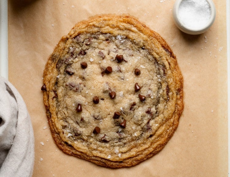 Large single-serve thin and chewy chocolate chip cookie on parchment paper with flaky salt, chocolate chips and milk on tray.