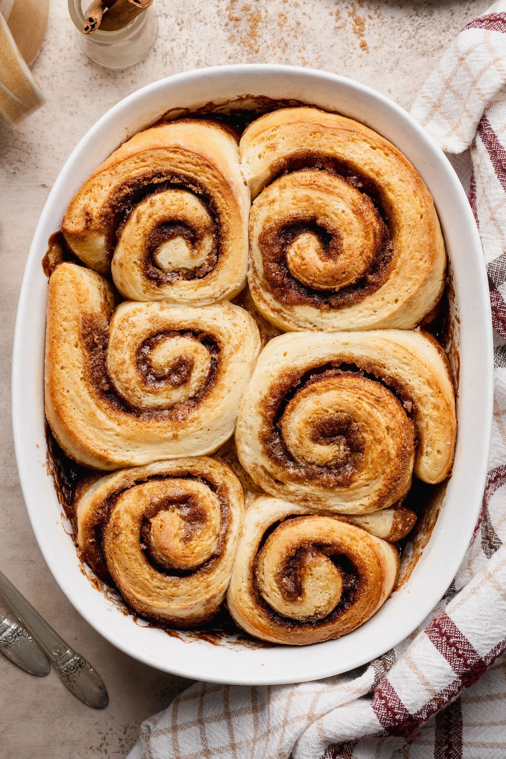 Overhead view of freshly baked cinnamon rolls in a white oval baking dish. The rolls are golden brown with visible cinnamon swirls and slightly caramelized edges.