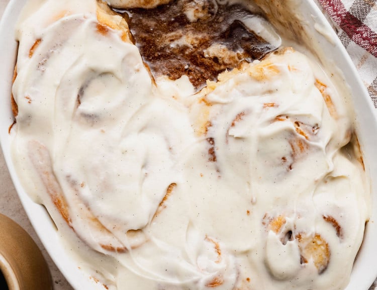 Overhead view of a baking dish with partially eaten cinnamon rolls covered in cream cheese frosting, revealing gooey cinnamon filling beneath.