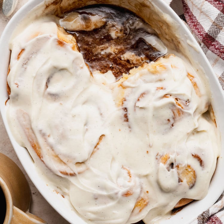 Overhead view of a baking dish with partially eaten cinnamon rolls covered in cream cheese frosting, revealing gooey cinnamon filling beneath.
