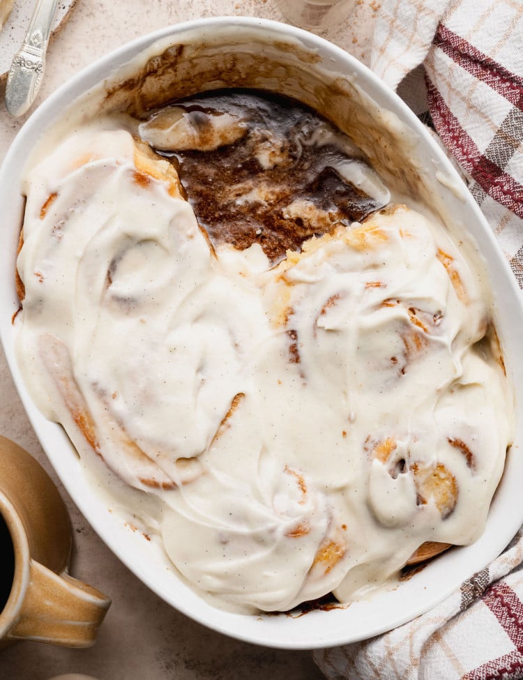 Overhead view of a baking dish with partially eaten cinnamon rolls covered in cream cheese frosting, revealing gooey cinnamon filling beneath.