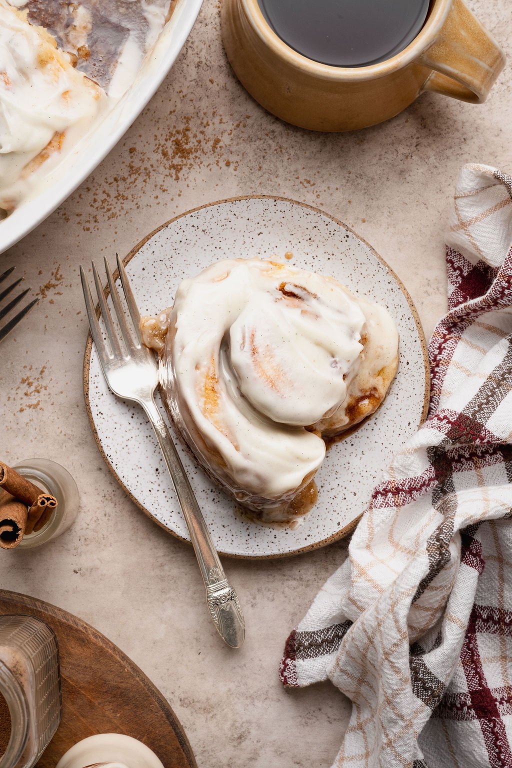 Overhead image of a frosted cinnamon roll served on a speckled plate with a fork beside a mug of coffee and a cozy plaid napkin.