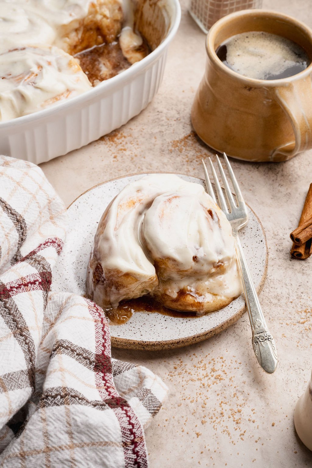Side angle of a frosted cinnamon roll on a plate with coffee in the background and a soft plaid napkin, creating a warm breakfast scene.
