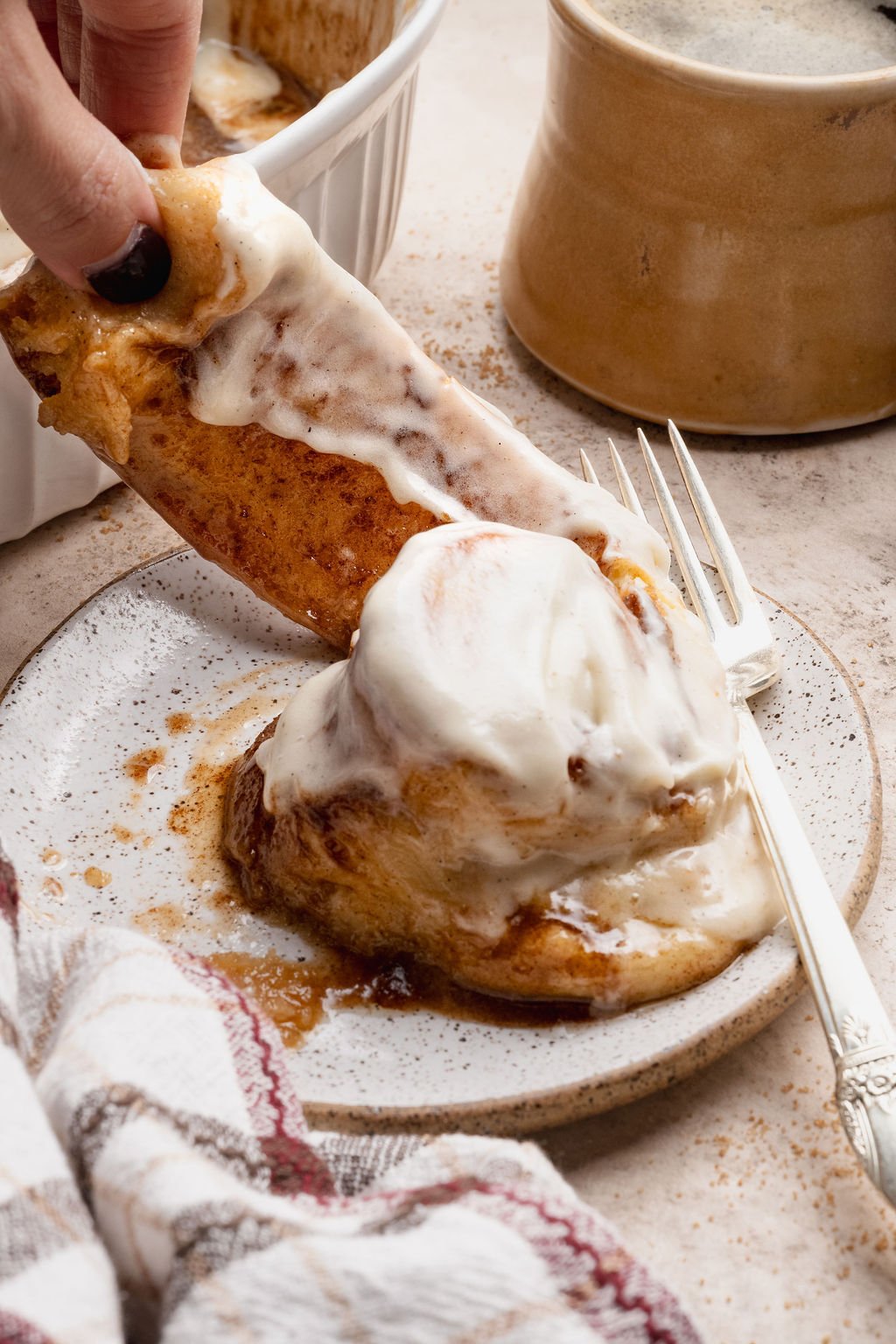 Close-up of a frosted cinnamon roll on a plate with a hand pulling apart a gooey, frosting-covered piece to show the soft interior.