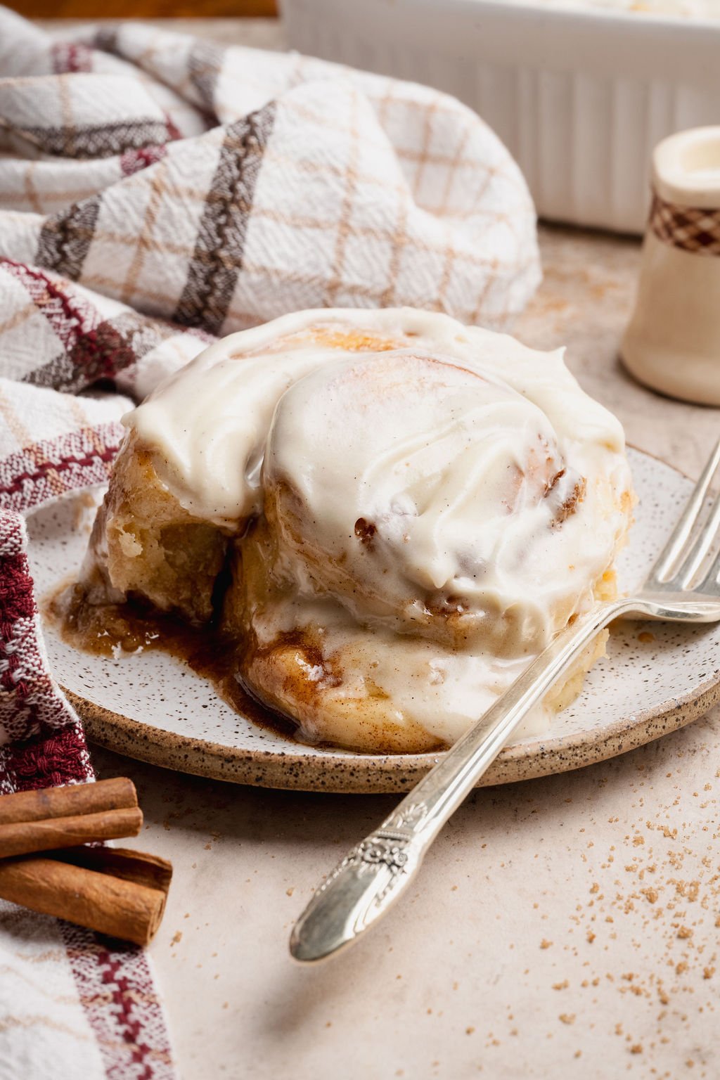 Close-up side view of a soft cinnamon roll topped with thick cream cheese frosting on a plate, surrounded by cozy breakfast props.