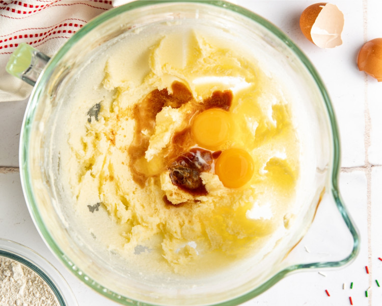 Creamed butter and sugar in a glass bowl with eggs and vanilla extract added on top, ready to be mixed for the cookie dough.