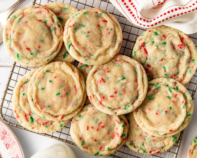 Baked Sugar Cookies with sprinkles cooling on a wire rack, with festive red and green sprinkles and a white napkin with red stitching beside them.