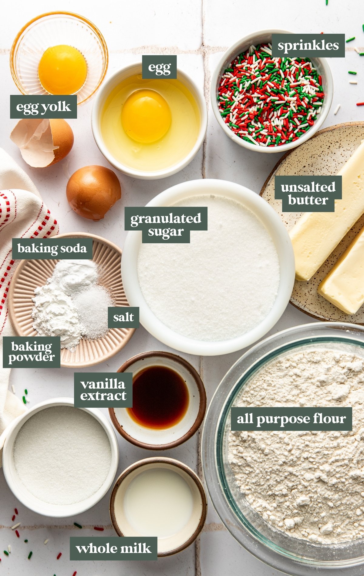 Overhead view of labeled ingredients for Christmas Sprinkle Sugar Cookies, including butter, sugar, egg, egg yolk, flour, baking powder, baking soda, salt, vanilla extract, milk, and red and green sprinkles arranged neatly on a white surface.