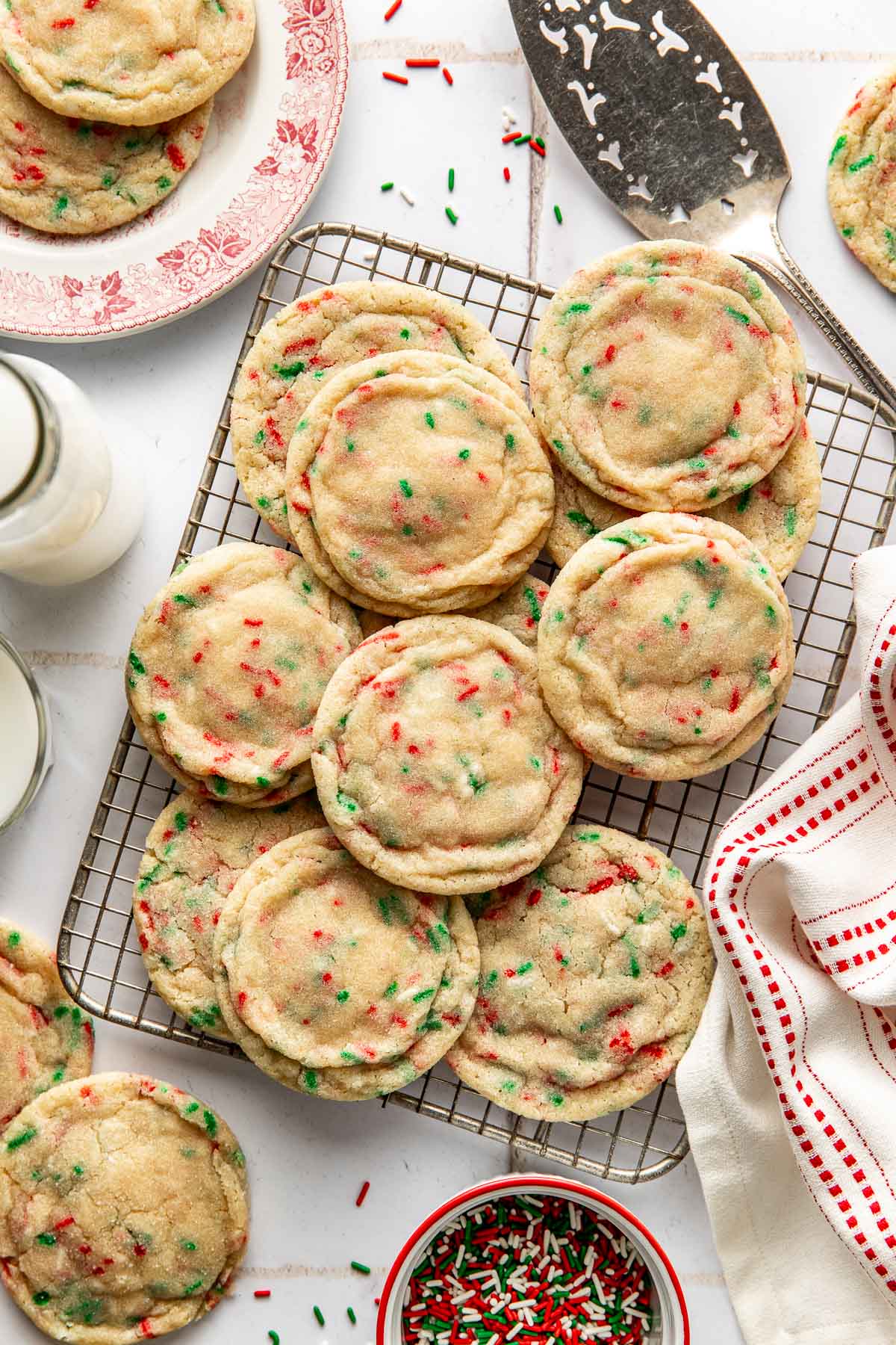 A cooling rack filled with soft and chewy Christmas sprinkle sugar cookies, speckled with red and green sprinkles, surrounded by a glass of milk, a bowl of extra sprinkles, and a festive red and white napkin.