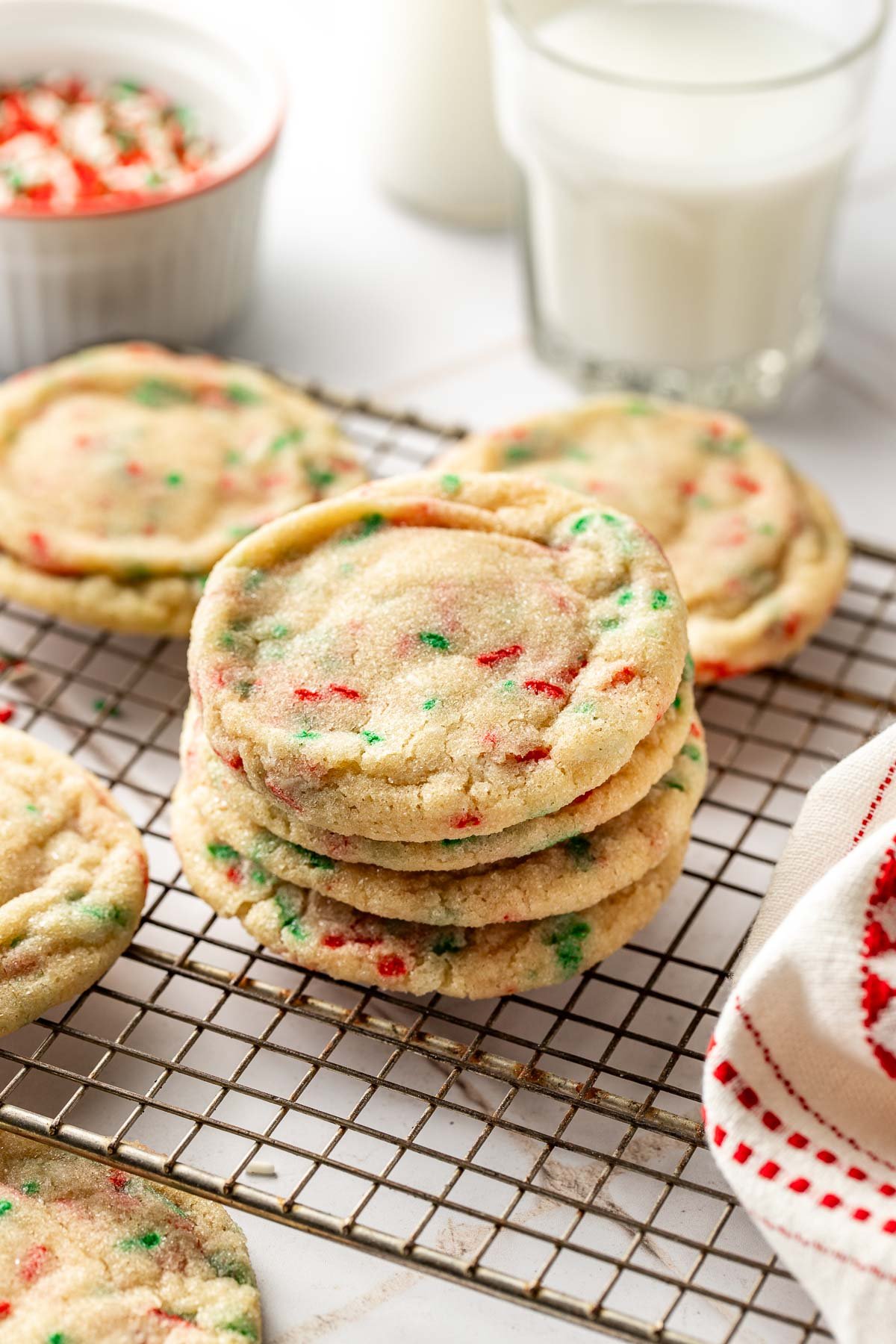 Stack of Christmas sprinkle sugar cookies on a cooling rack next to glasses of milk, with red and green sprinkles scattered around for a festive holiday touch.