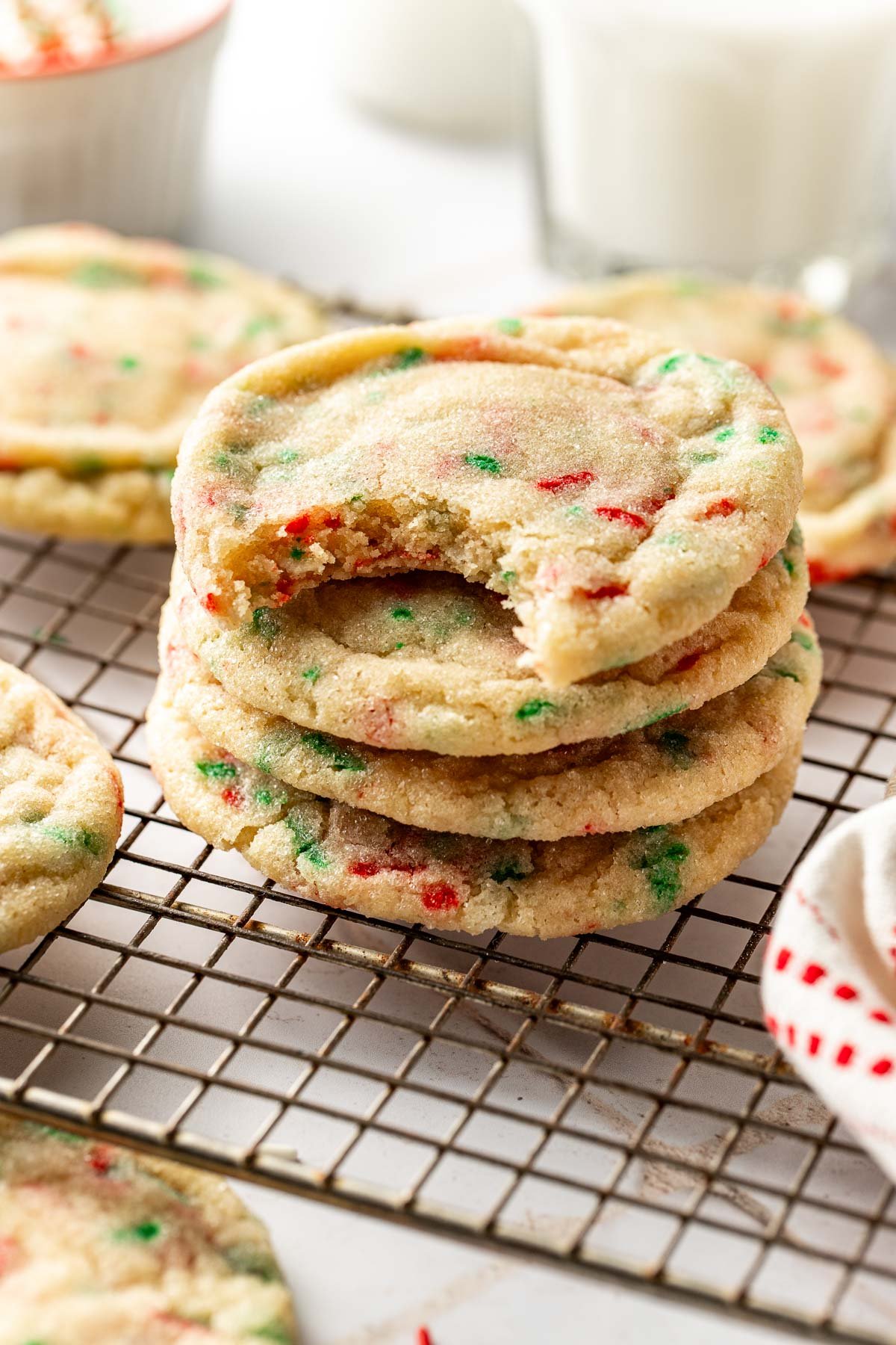 Stack of soft Christmas sugar cookies on a wire rack with one cookie missing a bite, showing their tender texture and colorful holiday sprinkles.