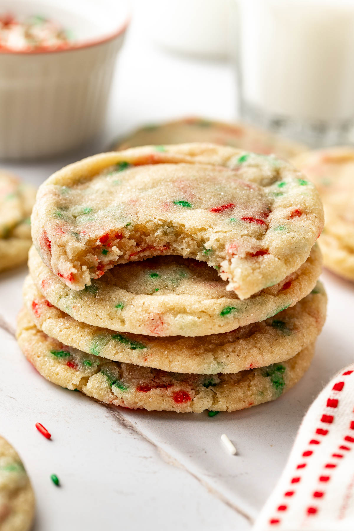 Side view of a stack of Christmas sprinkle sugar cookies with a bite taken out of the top cookie, set on a white surface with milk and a bowl of extra sprinkles in the background.