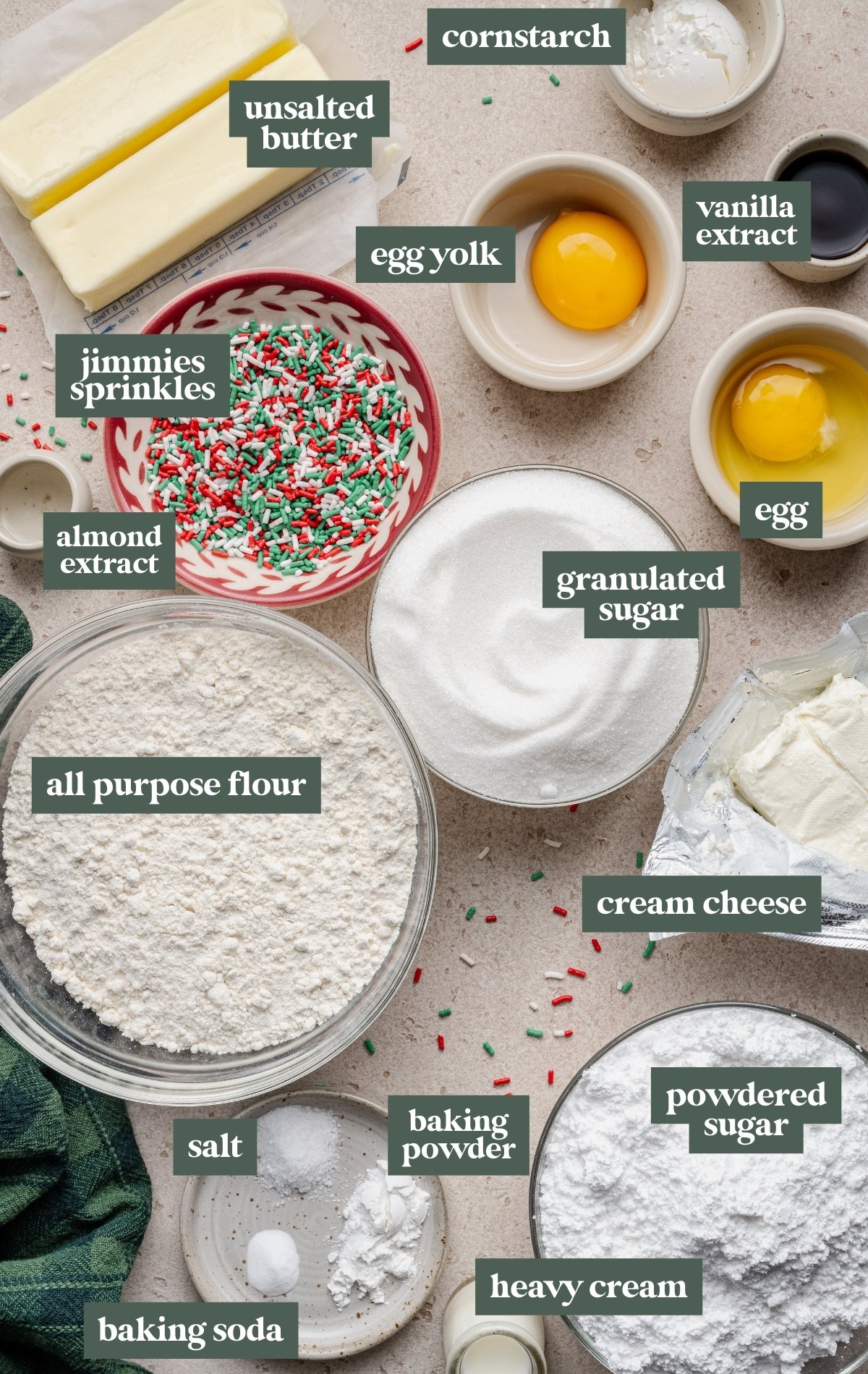 Overhead view of the ingredients for Christmas Sugar Cookie Bars arranged on a light countertop. Bowls and dishes hold all-purpose flour, granulated sugar, powdered sugar, jimmies sprinkles, cream cheese, cornstarch, baking soda, baking powder, salt, heavy cream, vanilla extract, almond extract, an egg, an egg yolk, and two sticks of unsalted butter. Festive red, green, and white sprinkles are scattered around the ingredients.