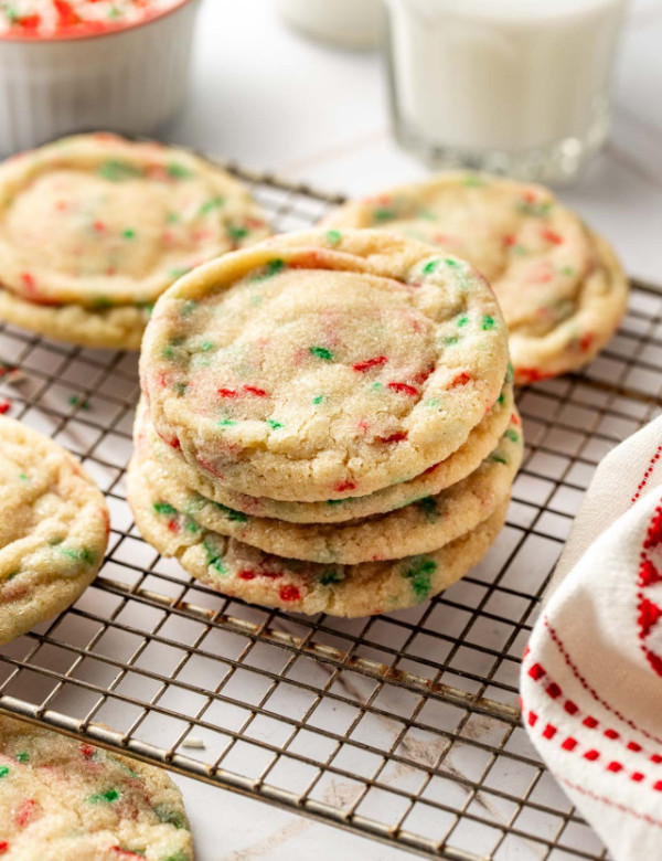 Stack of Christmas sprinkle sugar cookies on a cooling rack next to glasses of milk, with red and green sprinkles scattered around for a festive holiday touch.