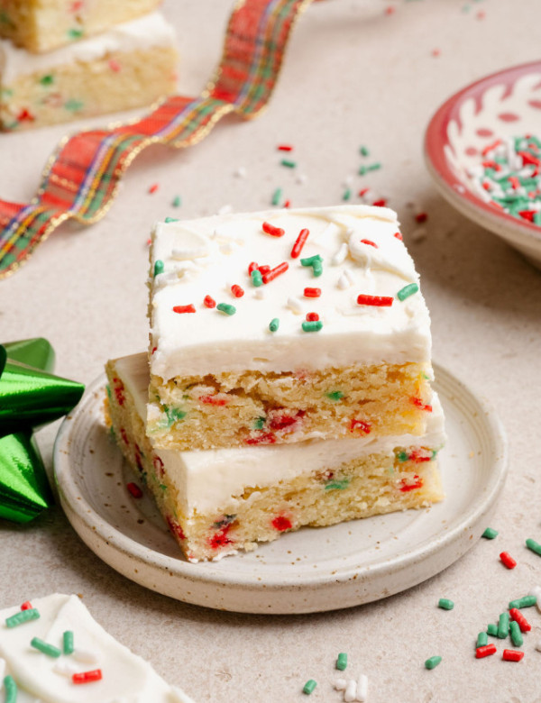 Two stacked Christmas sugar cookie bars on a small plate, showing a soft, sprinkle-studded interior and creamy white frosting with holiday sprinkles.