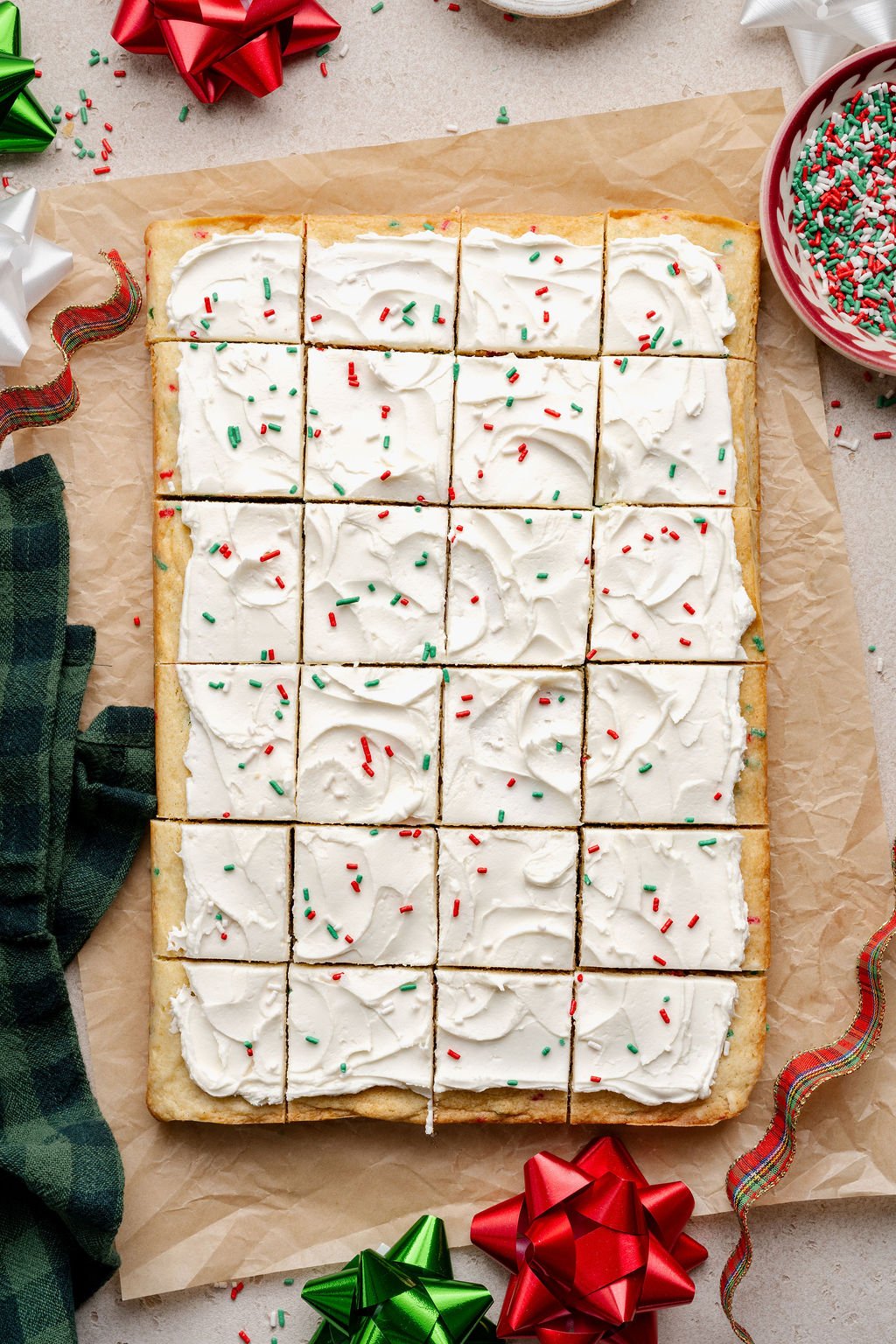 A full pan of Christmas sugar cookie bars cut into even squares, each topped with swirled white frosting and holiday sprinkles, with bowls of sprinkles and gift bows surrounding the scene.