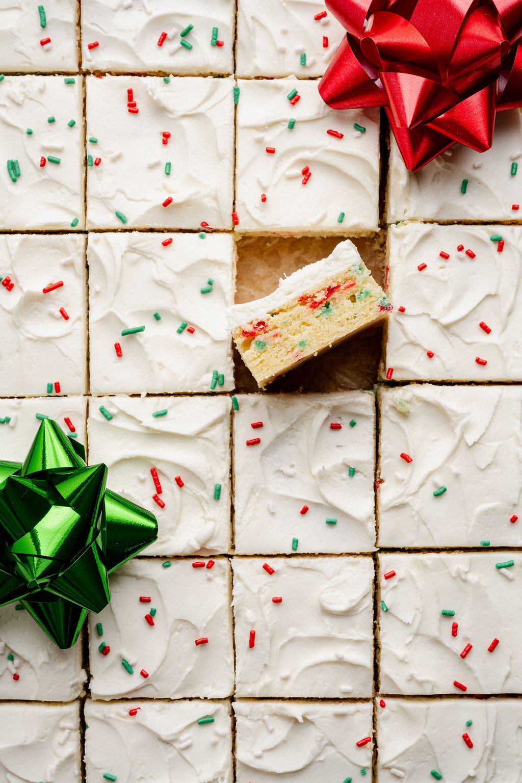 A grid of frosted Christmas sugar cookie bars with one bar lifted slightly to show the festive red and green sprinkles baked inside, surrounded by holiday bows.