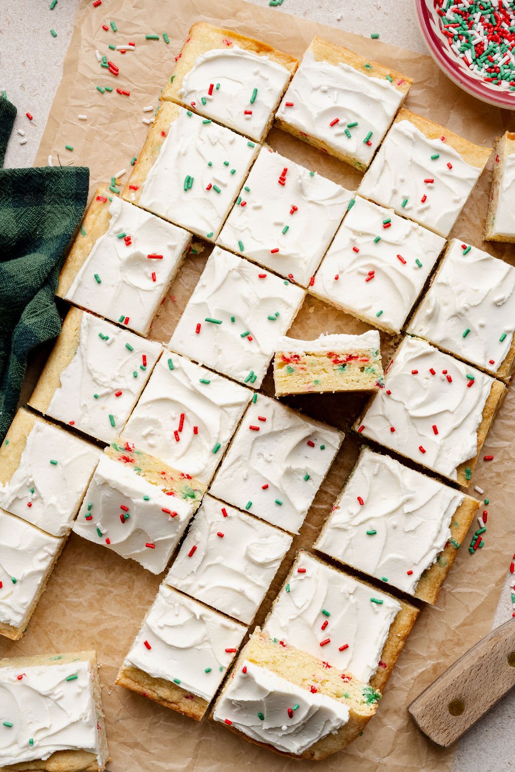Christmas sugar cookie bars arranged on parchment paper, with one bar pulled out to reveal the soft interior full of red and green sprinkles and topped with thick white frosting.