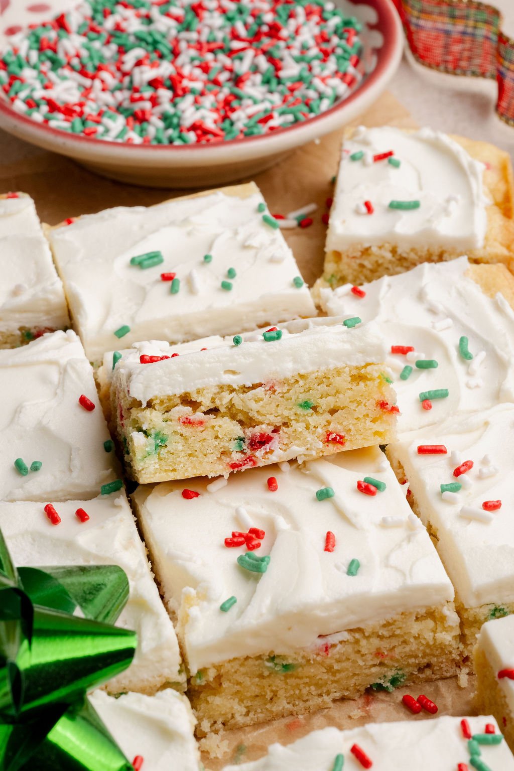 A close-up of Christmas sugar cookie bars topped with swirled white frosting and festive sprinkles, with a bowl of sprinkles and holiday ribbon in the background.