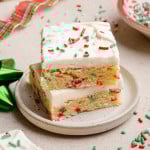 Two stacked Christmas sugar cookie bars on a small plate, showing a soft, sprinkle-studded interior and creamy white frosting with holiday sprinkles.
