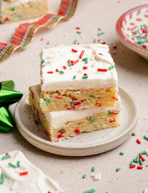 Two stacked Christmas sugar cookie bars on a small plate, showing a soft, sprinkle-studded interior and creamy white frosting with holiday sprinkles.