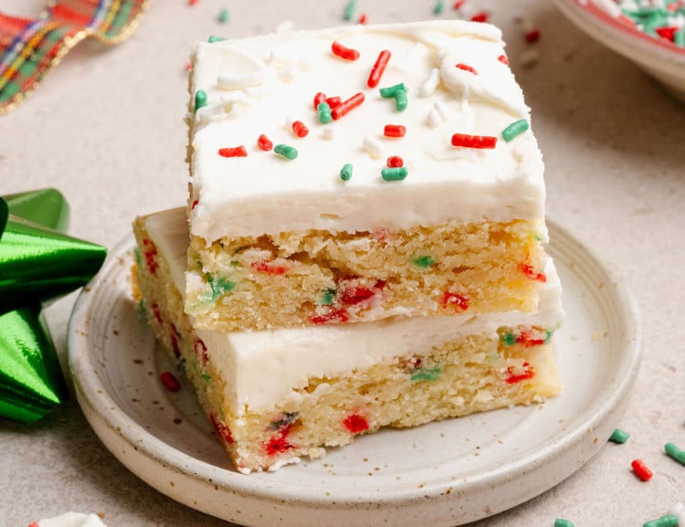 Two stacked Christmas sugar cookie bars on a small plate, showing a soft, sprinkle-studded interior and creamy white frosting with holiday sprinkles.