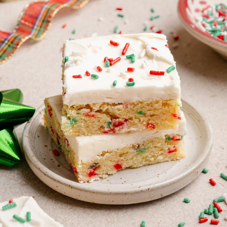 Two stacked Christmas sugar cookie bars on a small plate, showing a soft, sprinkle-studded interior and creamy white frosting with holiday sprinkles.