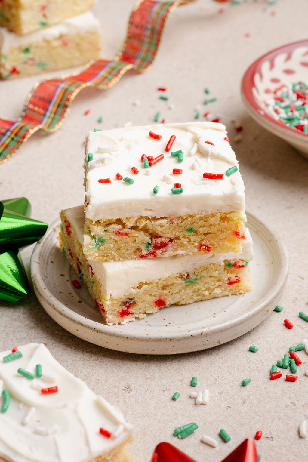 Two stacked Christmas sugar cookie bars on a small plate, showing a soft, sprinkle-studded interior and creamy white frosting with holiday sprinkles.