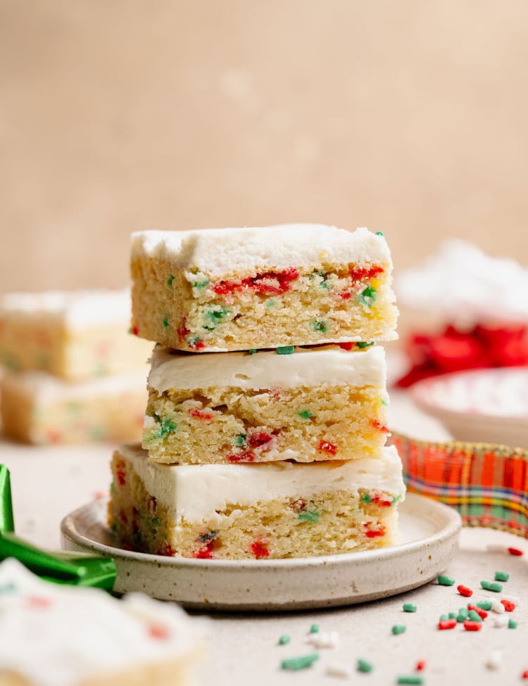A tall stack of three Christmas sugar cookie bars on a small plate, highlighting their soft texture and colorful sprinkles baked into the cookie layer.