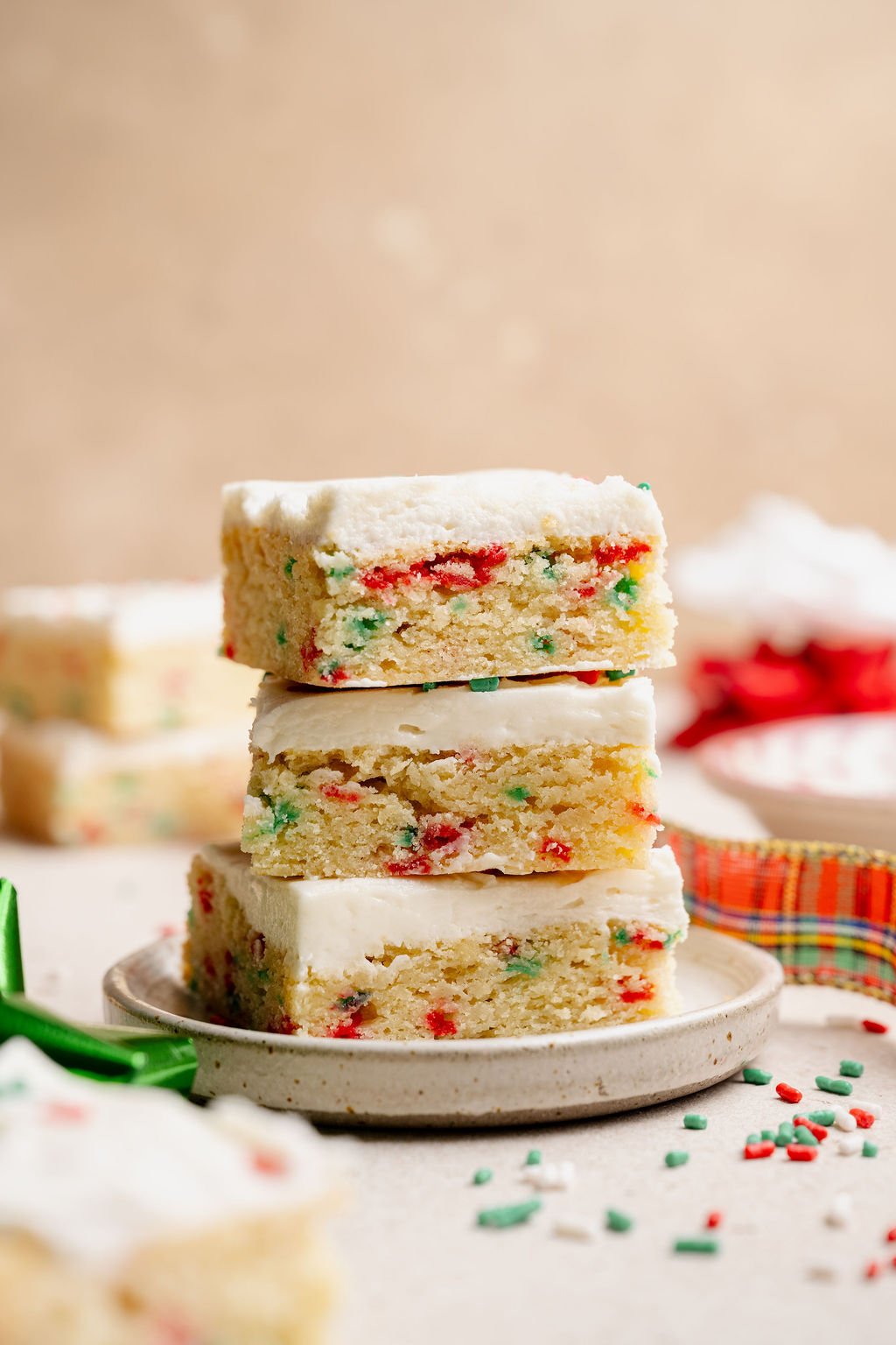 A tall stack of three Christmas sugar cookie bars on a small plate, highlighting their soft texture and colorful sprinkles baked into the cookie layer.