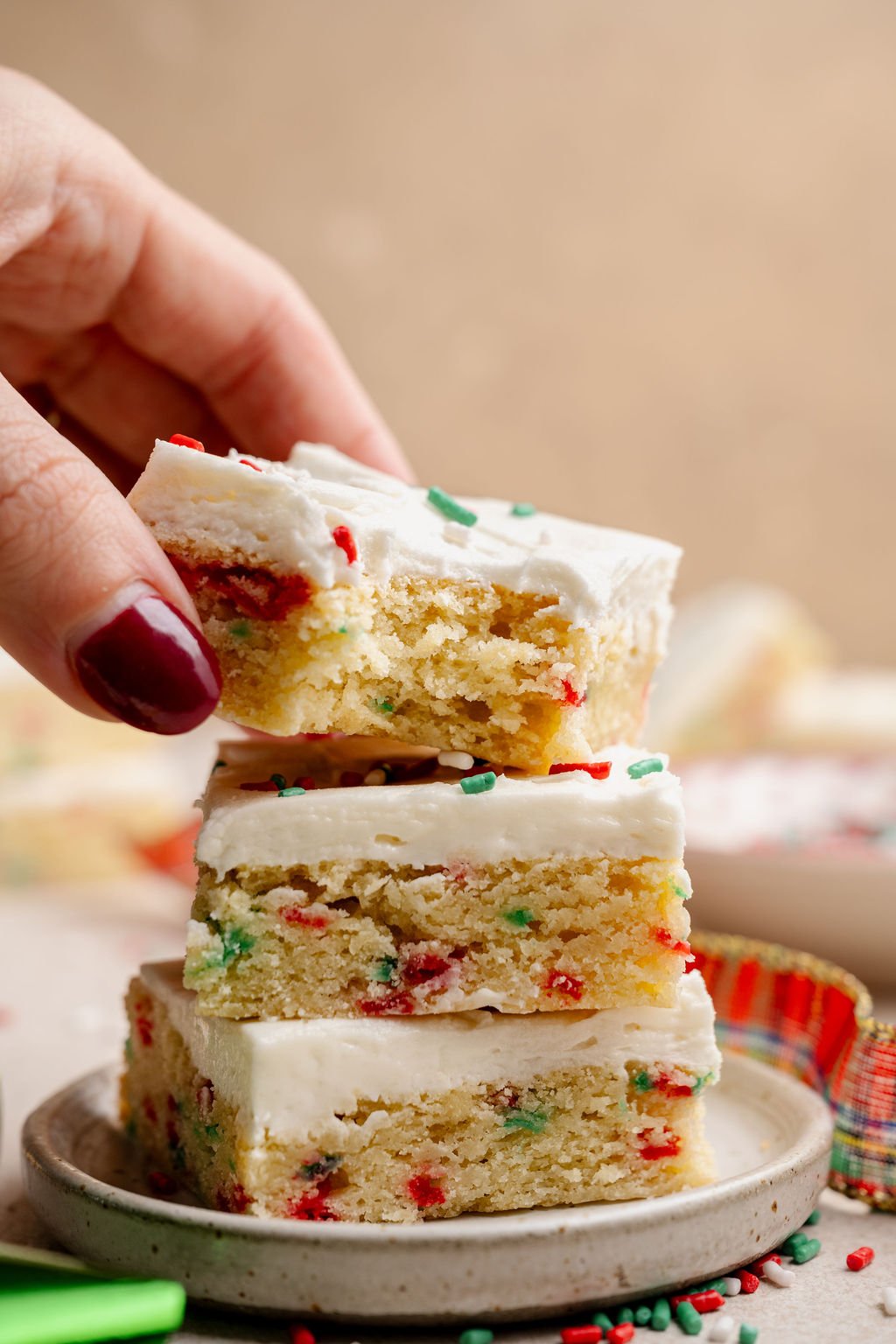 A hand lifting a frosted Christmas sugar cookie bar from a stack, showcasing the moist, sprinkle-filled interior and thick layer of white frosting.