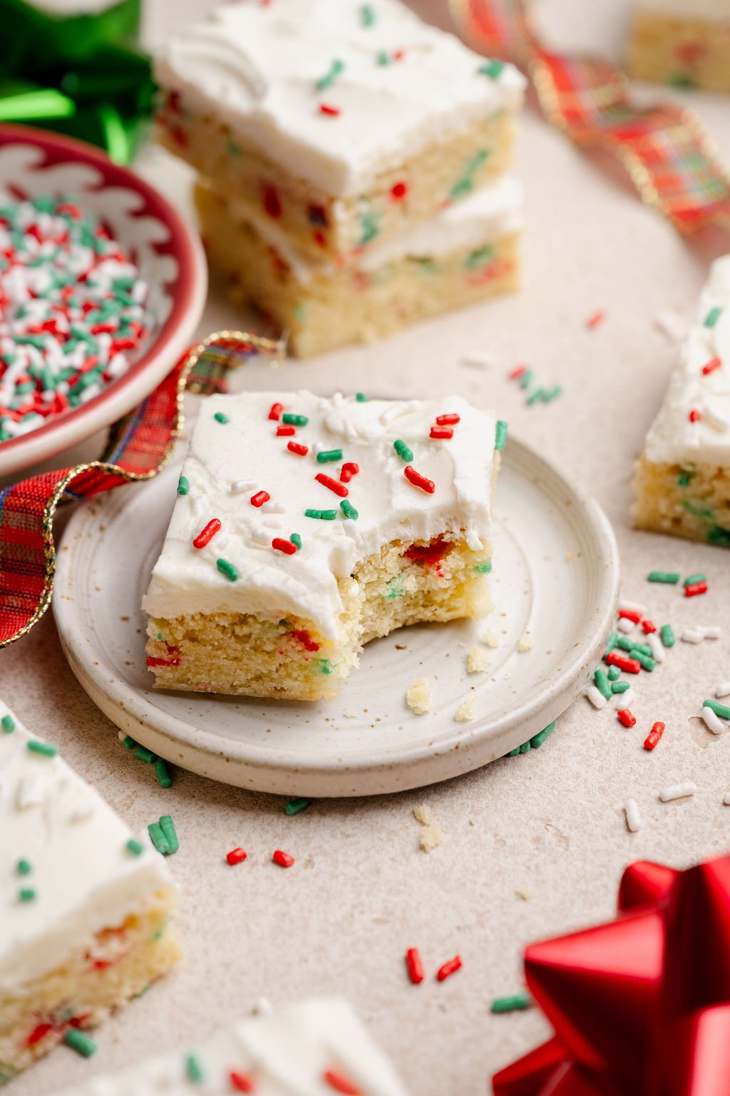 A Christmas sugar cookie bar on a small plate with a bite taken out, surrounded by scattered holiday sprinkles and festive ribbon.