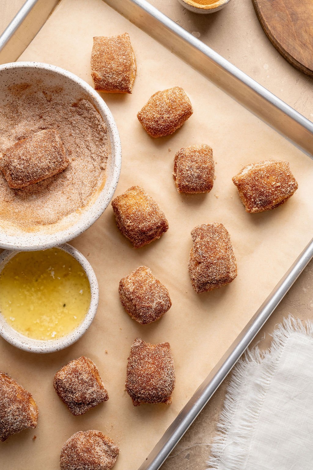 Overhead view of cinnamon sugar pretzel bites arranged on a parchment-lined baking sheet with bowls of melted butter and cinnamon sugar nearby.