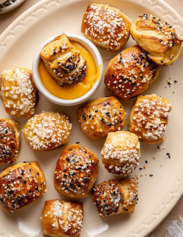 Overhead view of soft, golden-brown pretzel bites on a cream-colored plate, topped with pretzel salt and everything bagel seasoning, with one bite dipped into a bowl of warm cheese sauce in the center.