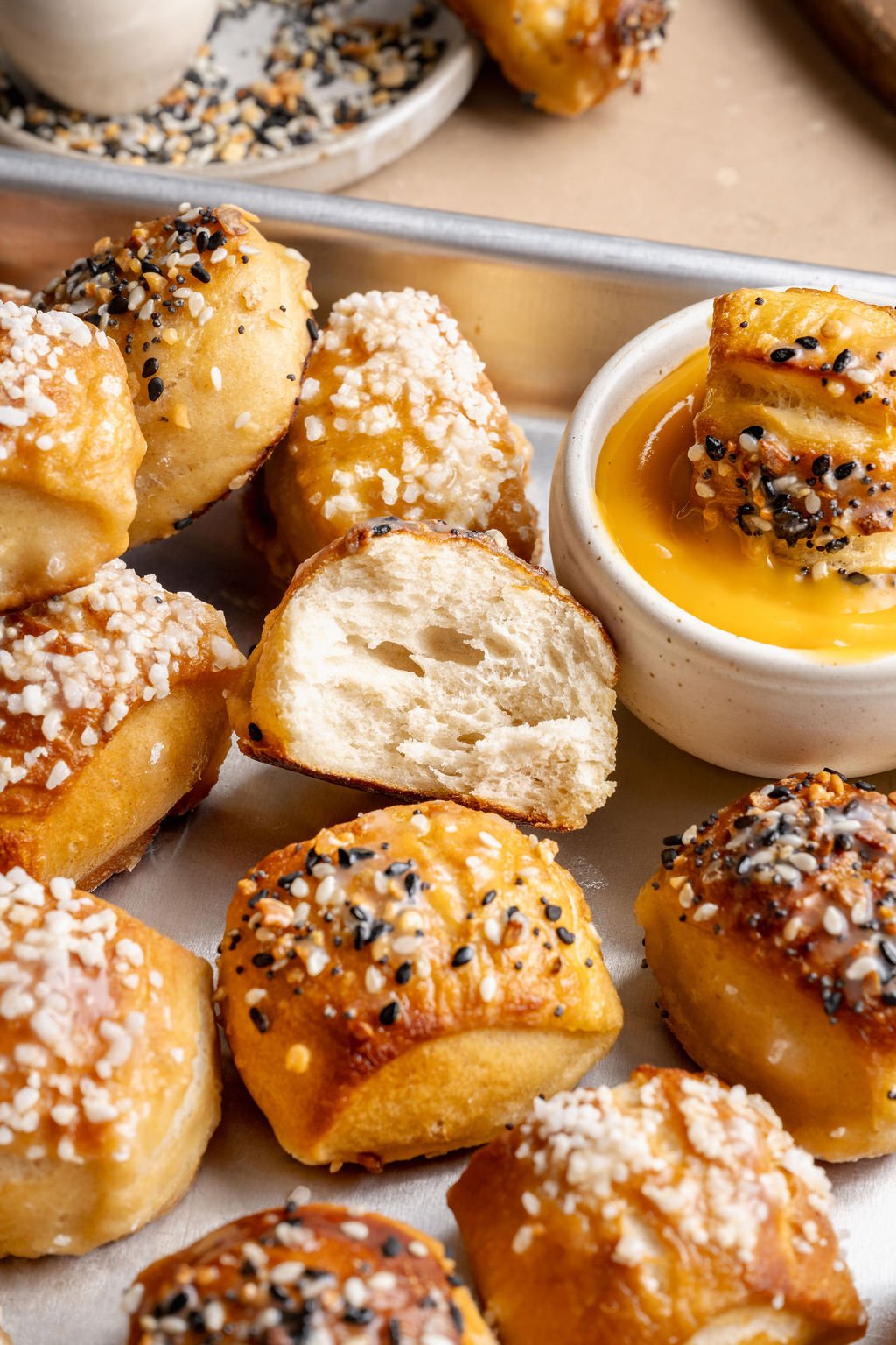 Close-up view of soft pretzel bites on a metal tray with a bite broken open to show the fluffy interior next to cheese sauce.