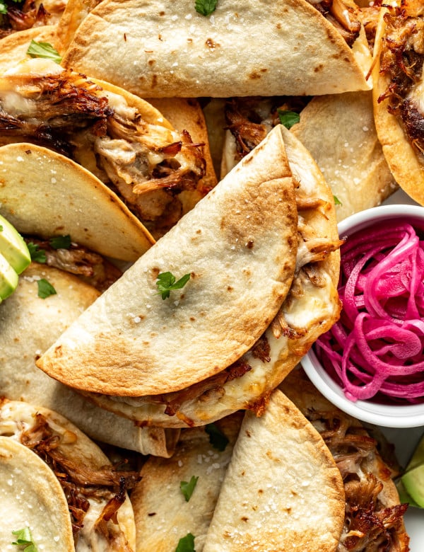 Crispy baked pork tacos piled on top of each other with small bowl of pickled red onions on the side.