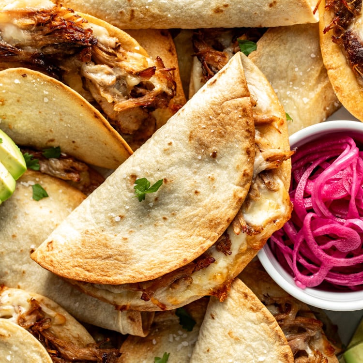 Crispy baked pork tacos piled on top of each other with small bowl of pickled red onions on the side.