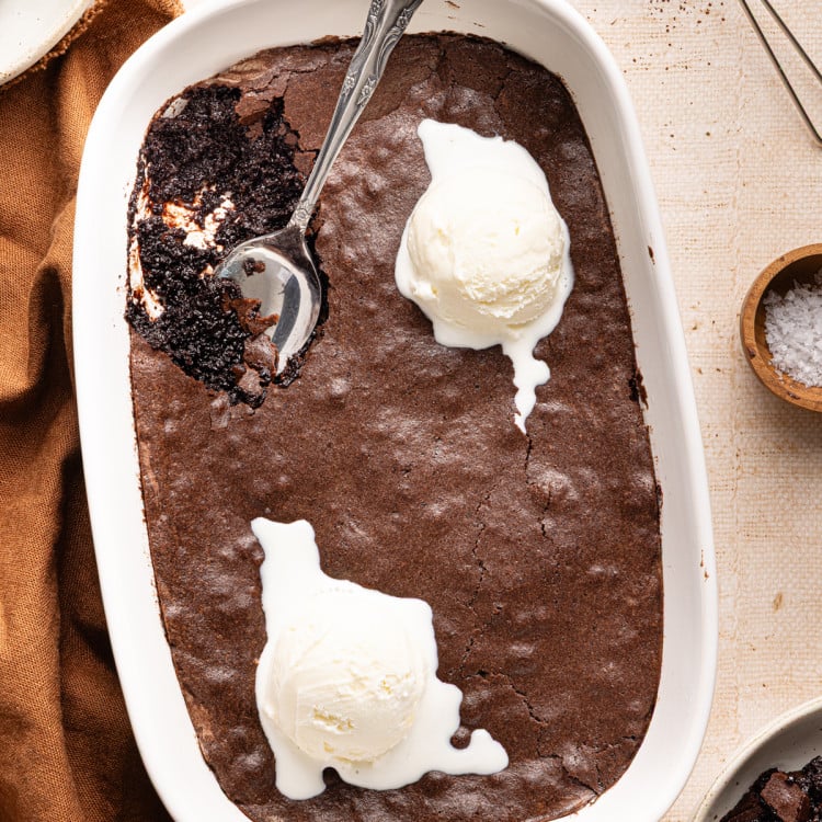 Brownie pudding in baking dish with two scoops of vanilla ice cream and spoon in top left corner.
