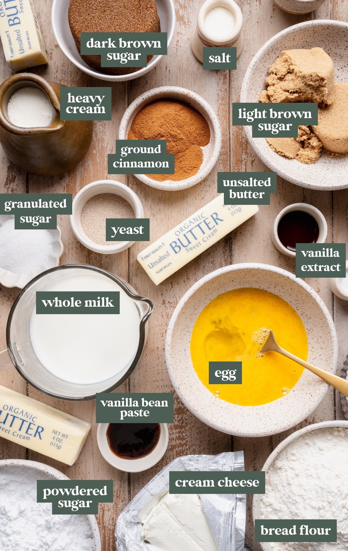 Overhead flat lay of ingredients for overnight cinnamon rolls including bread flour, yeast, whole milk, eggs, butter, cream cheese, cinnamon, brown sugar, granulated sugar, powdered sugar, vanilla extract, vanilla bean paste, heavy cream, and salt, each labeled on a neutral surface.