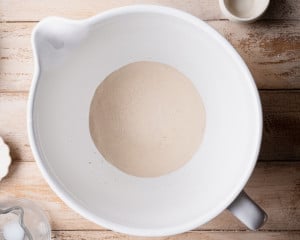 Overhead view of active dry yeast blooming in warm milk inside a white mixing bowl, showing foamy bubbles forming on the surface.